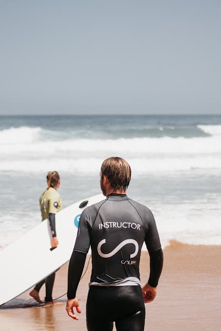 A surf instructor and student holding his surfboard looking at the waves.